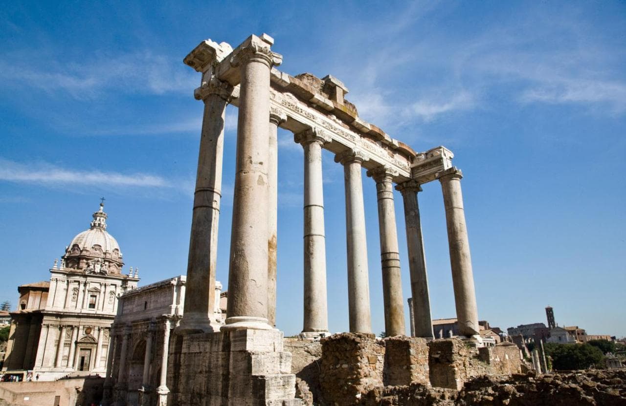 The standing columns of a ruined ancient temple against a blue sky, with a domed church behind them.